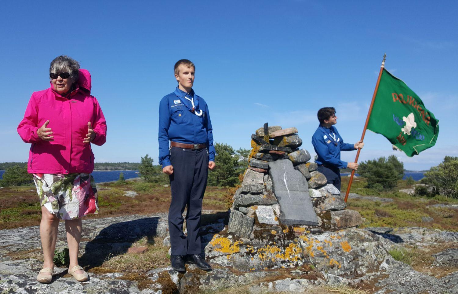 Kvinna och två ynglingar står vid monument på klippa