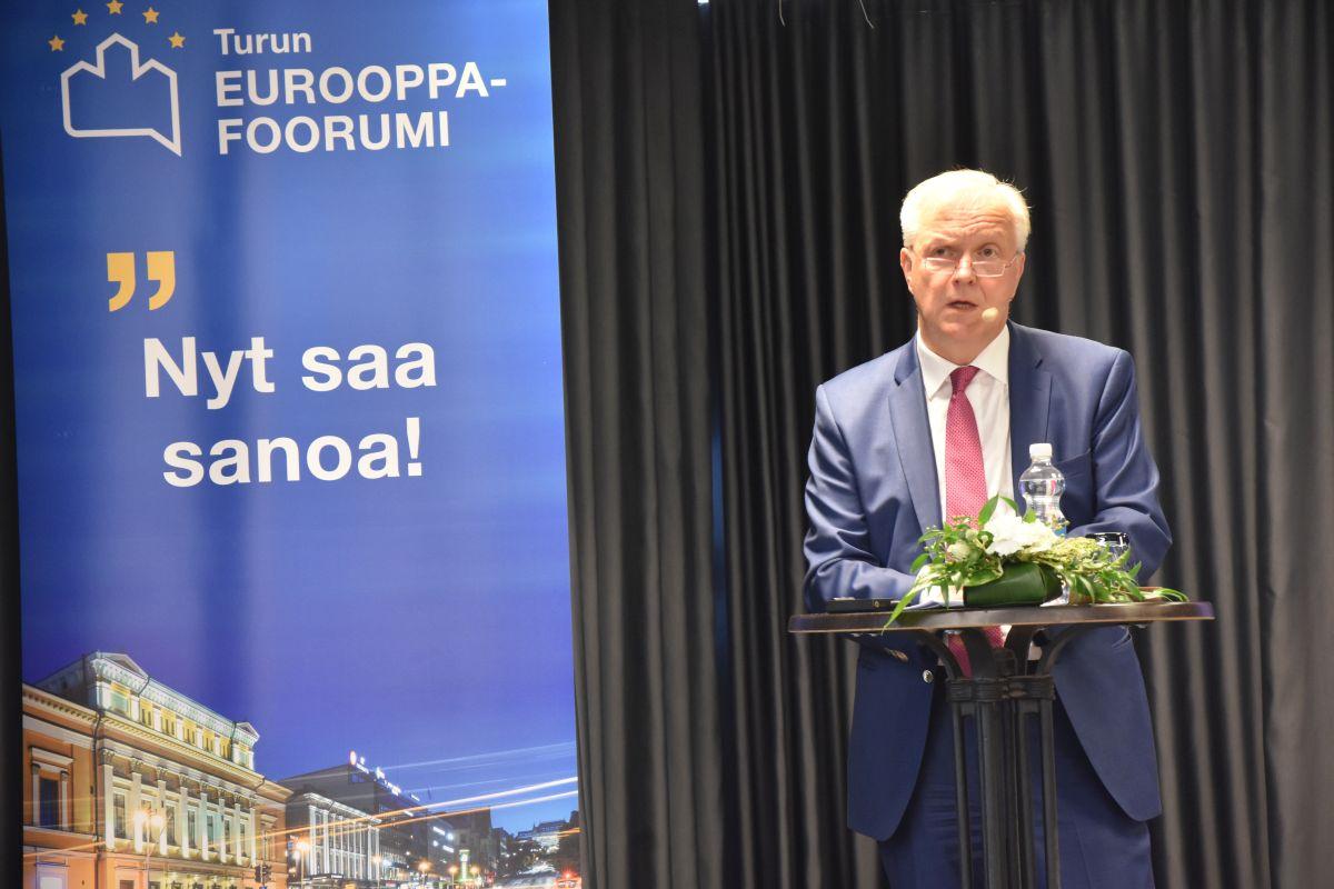 Olli Rehn står på en scen och talar.