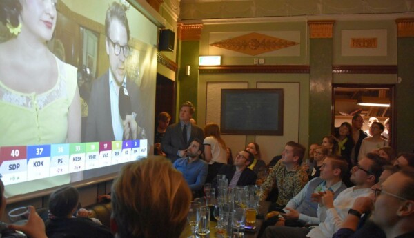 Fullsatt barsal med många människor som tittar på en stor skärm med tv-program.