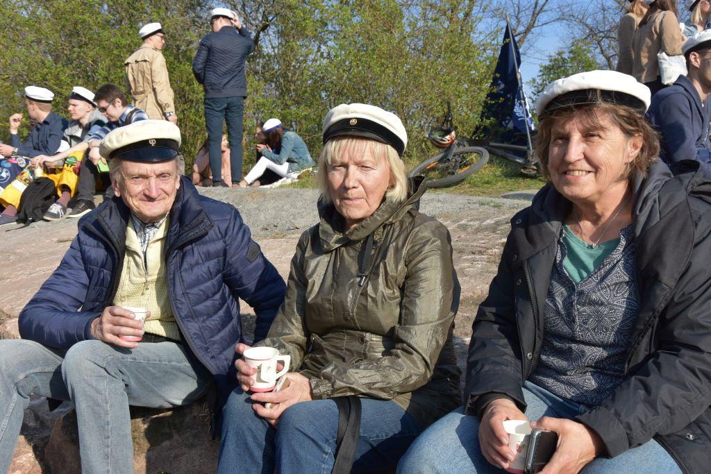 Tre personer iklädda studentmössor sitter på ett berg i en park. I bakgrunden ser man ungdomar som firar.