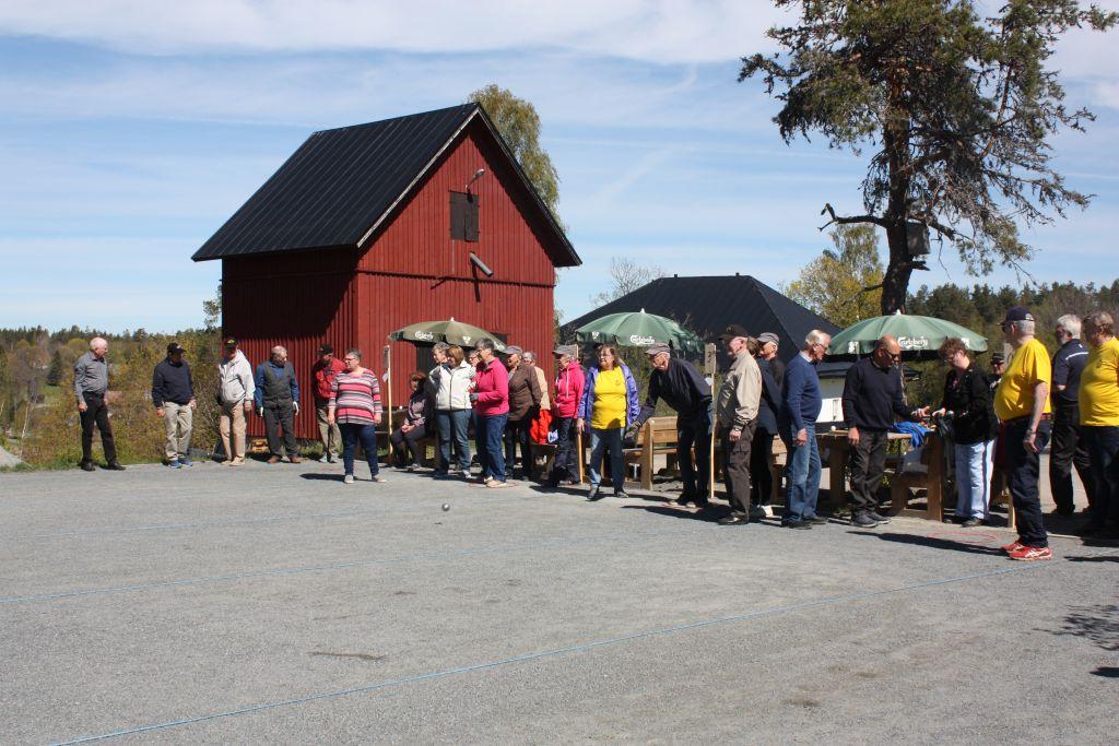 Sandplan med personer i bakgrunden som står uppradade längs med en linje utomhus, blå himmel, färggranna kläder, rött hus i bakgrunden.