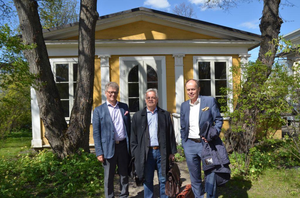 Tre män klädda i kostym står framför ett gult lusthus omringade av växtlighet.