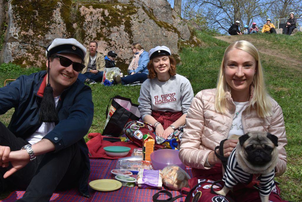 Tre personer och en hund sitter på en picknickfilt i en park