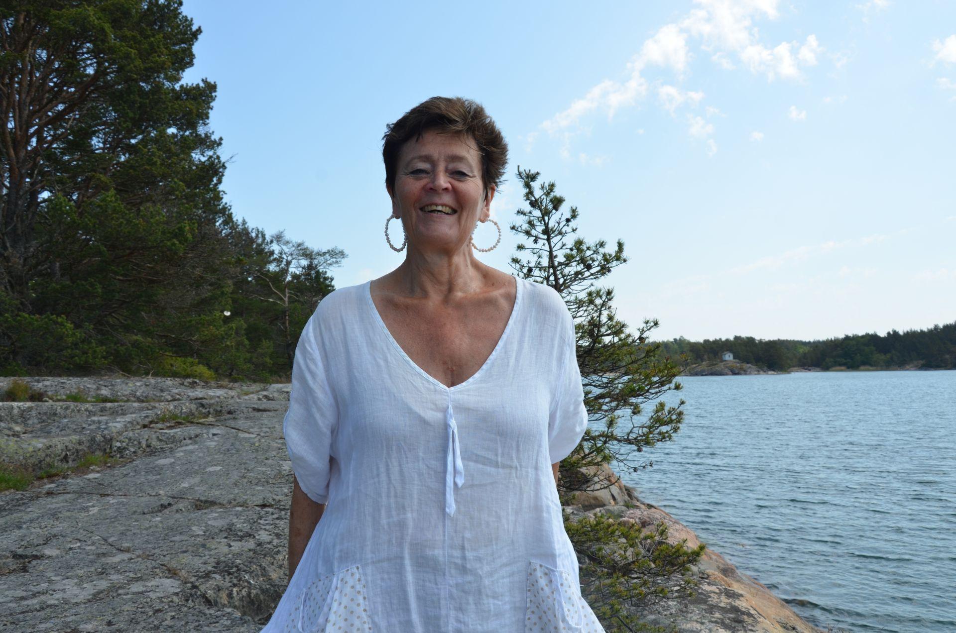 En kvinna står och skrattar på ett berg. I bakgrunden ser man havet och barrträd.
