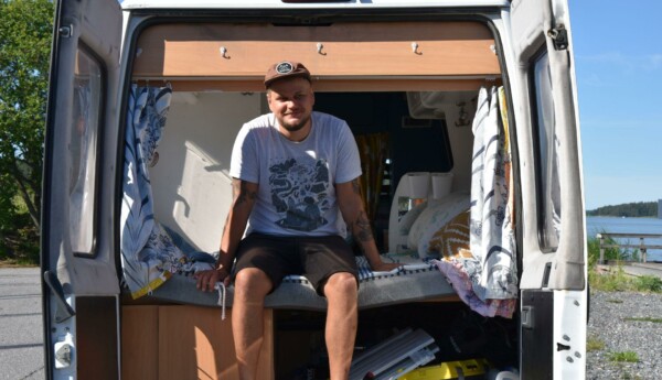 En man klädd i shorts, t-skjorta och keps sitter på en säng som finns i den öppna bakluckan av en paketbil.