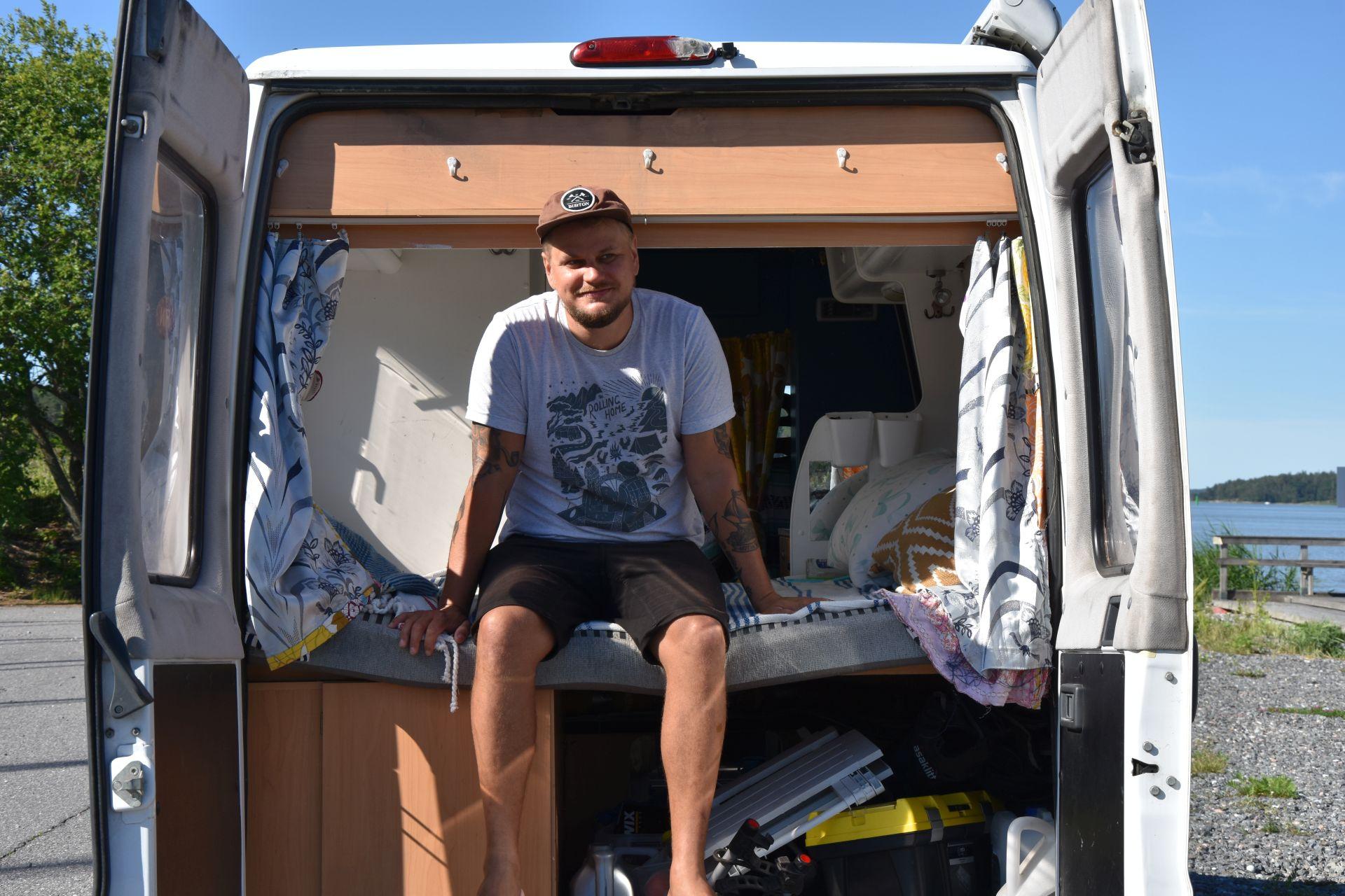 En man klädd i shorts, t-skjorta och keps sitter på en säng som finns i den öppna bakluckan av en paketbil.