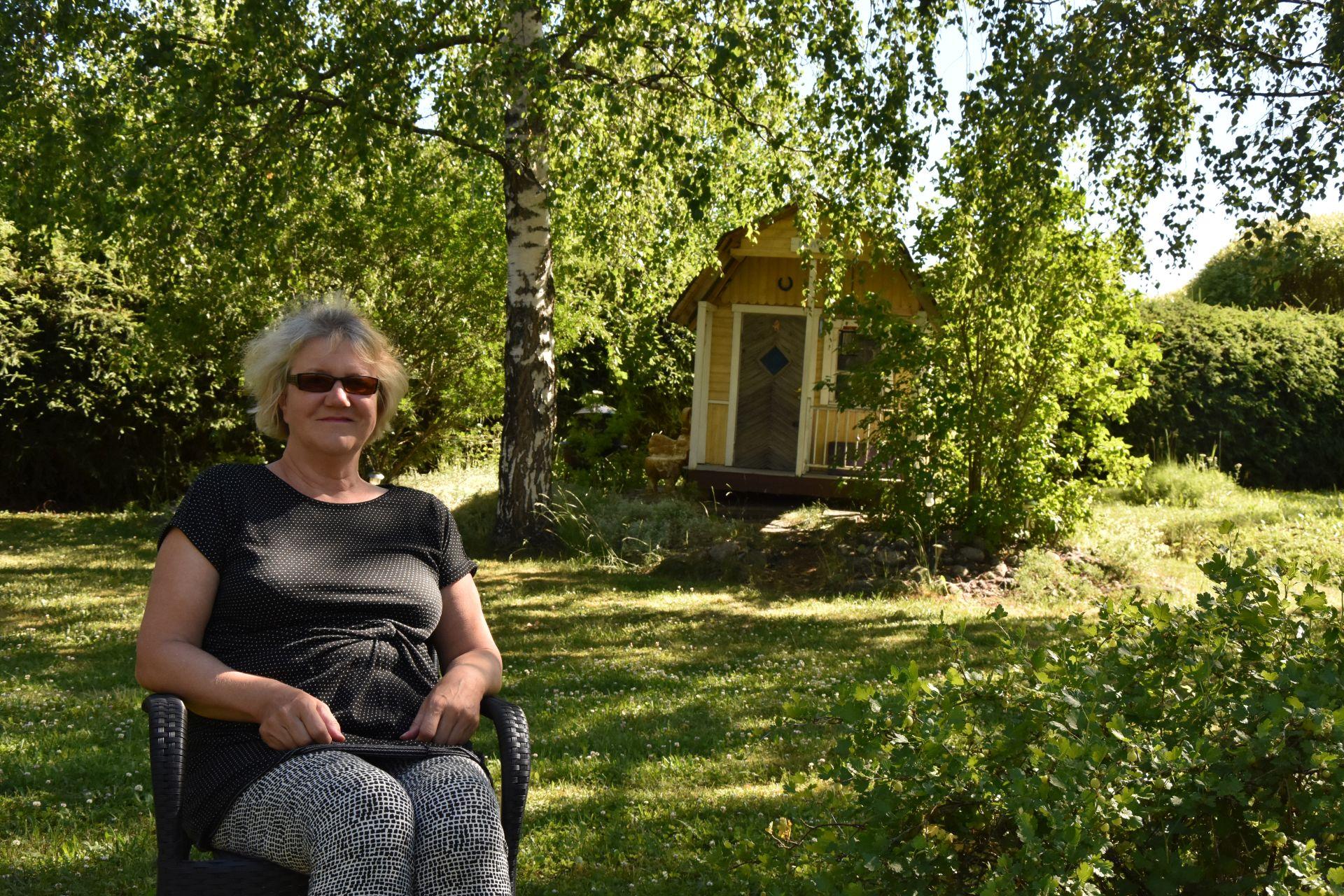 En kvinna sitter på en stol i en trädgård, i bakgrunden syns en gul lekstuga.