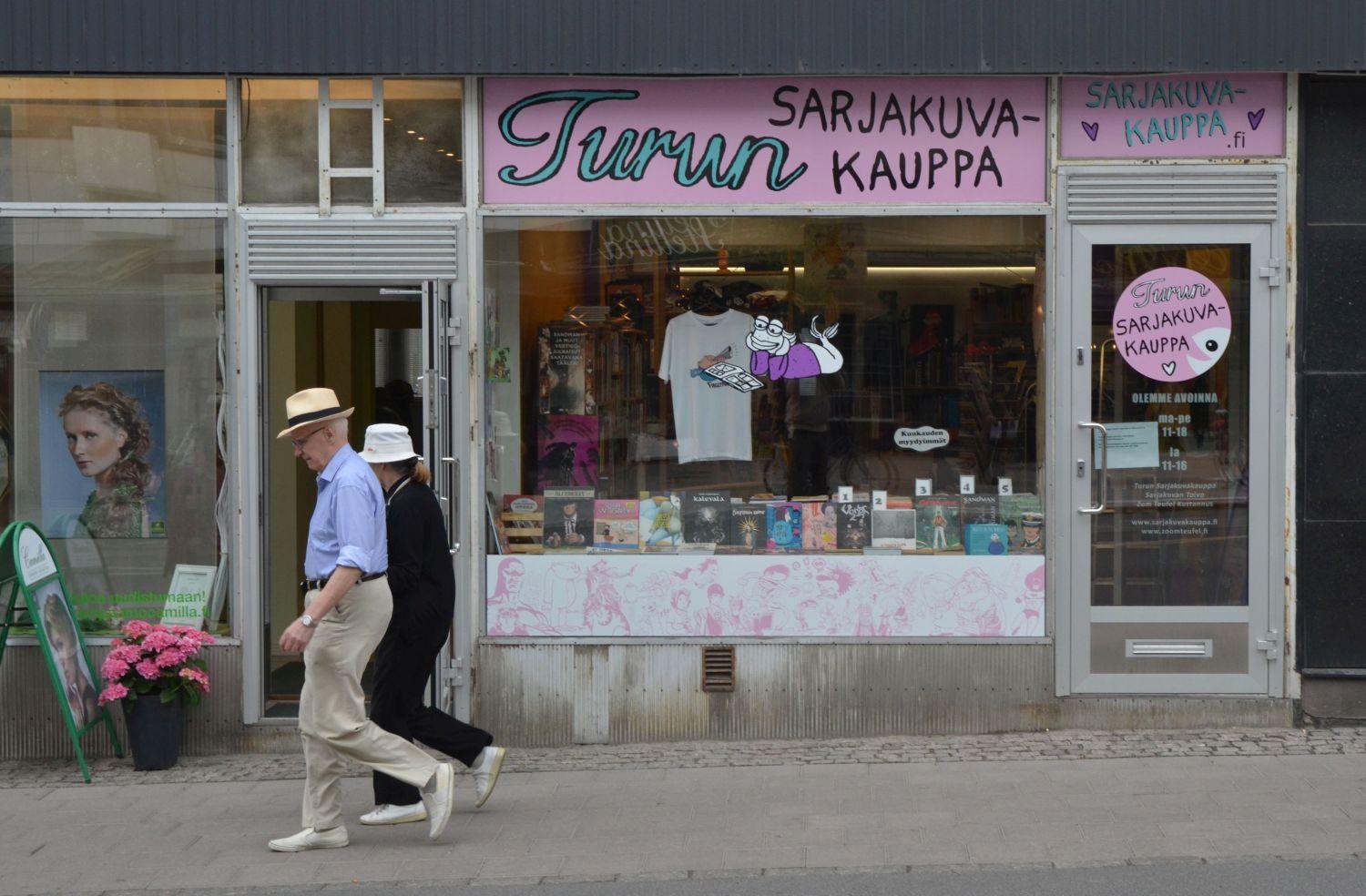 Två äldre människor går förbi en serieaffärs skyltförnster.