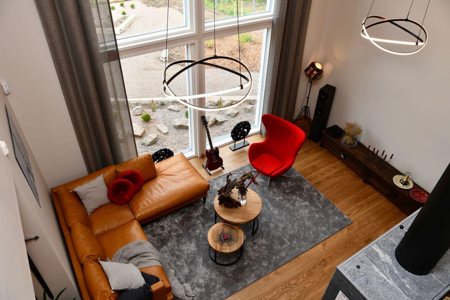 Vardagsrummet i Airon Neito fotograferat uppifrån. I vardagsrummet finns en ljusbrun soffa, en illröd fåtölj och en modern öppen spis. Fönstren i rummet är stora och det är högt i tak.