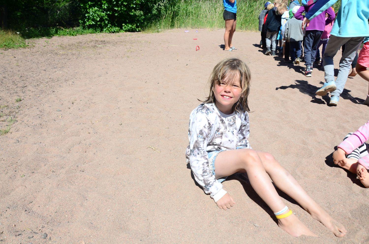Alicia Friman sitter på sandstranden. Bakom henne leker de andra barnen.