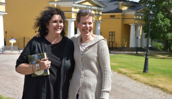 Två kvinnor står och håller om varandra utanför stora gula stenhus.