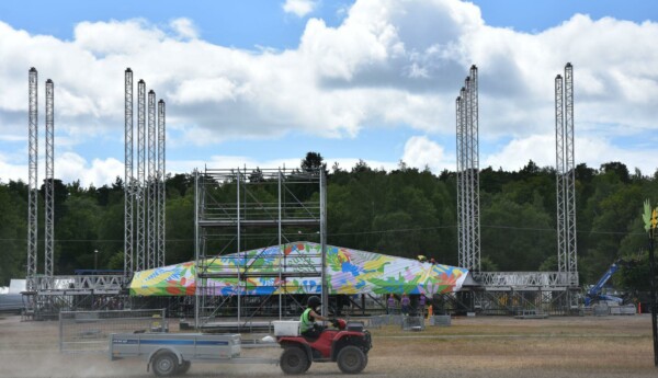 En festivalscen håller på att byggas. Scenen har fortfarande inget tak och framför scenen kör en person klädd i arbetskläder på en fyrhjuling.