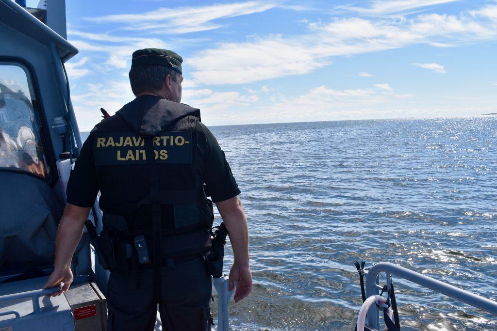 En sjöbevakare står på en båt och tittar ut över havet. På hans rygg står det "Rajavartiolaitos"