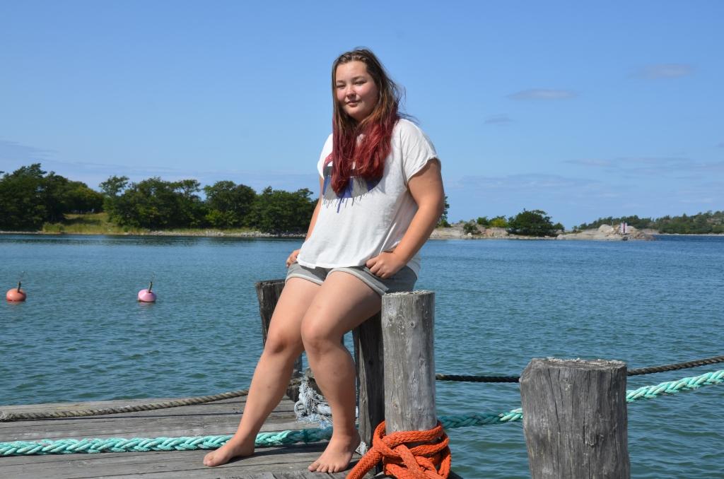 Emma Brunström sitter på en förtöjningspåle. I bakgrunden havet och delar av Tunhamn. Himlen är blå.