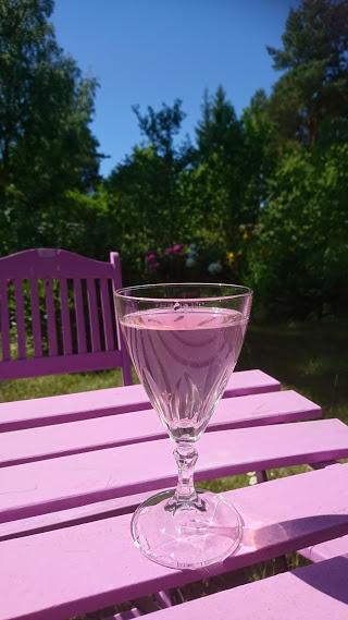 Ett glas med en rosa dryck står på ett rosa bord i en trädgård.