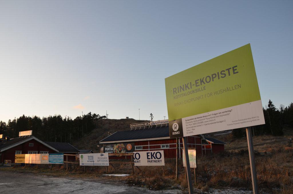 En skylt med texten "Rinki-ekopunkt för hushållen", bakom skylten syns Hirvensalos skidbacke.