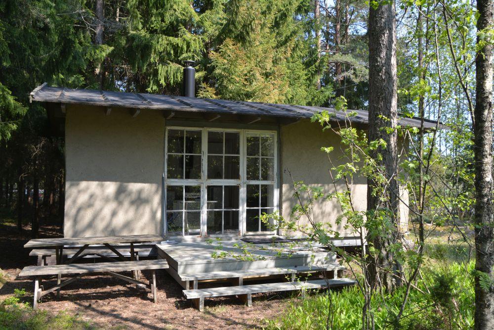 En liten vit stuga med ett stort fönster står i en skog.