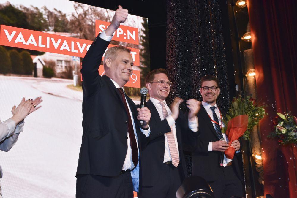 Tre kostymklädda och leende män på en scen. En man visar tummen upp och en annan klappar.