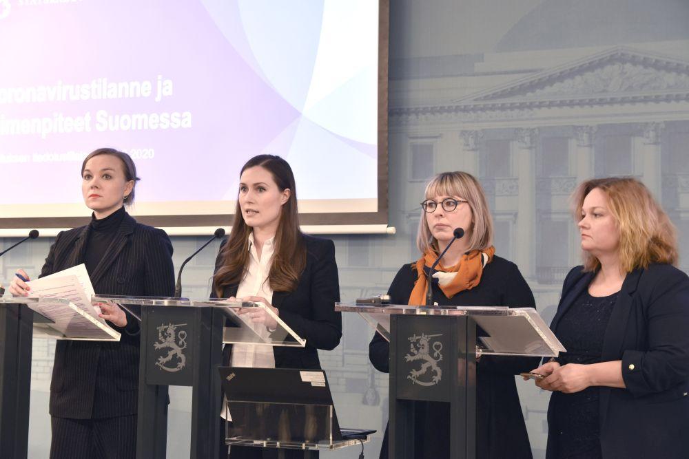 Fyra formellt klädda kvinnor står framför podium med mikrofoner och ser seriösa ut.