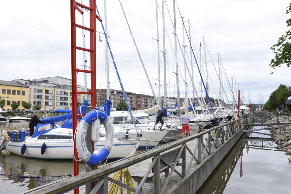 En rad med segelbåtar förtöjda vid en gästhamn. En person ombord på en av segelbåtarna räcker något till en person som står på bryggan.