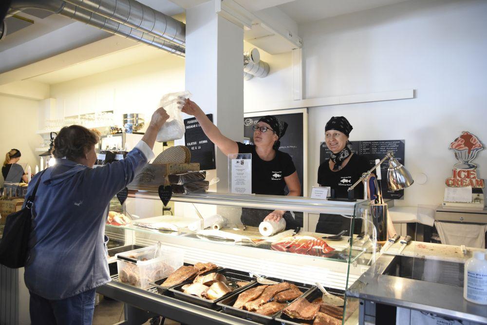 Två kvinnor klädda i svart t-skjorta och huvudduk står bakom en disk med fisk. En av kvinnorna räcker över en påse till en äldre dam på andra sidan disken.