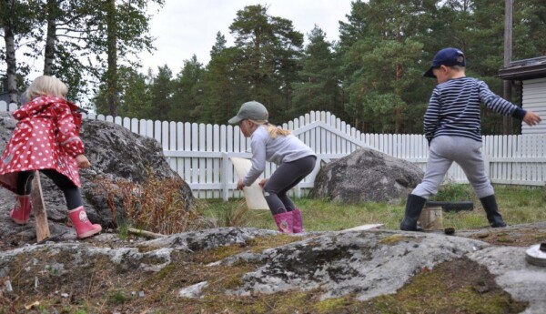 Tre barn leker med träbitar bland stenar på en bakgård, Daghemmet Karusellen i nagu