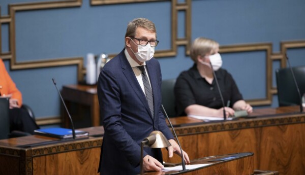 Kostymklädd herre i ansiktsmask vid riksdagens plenisals podium