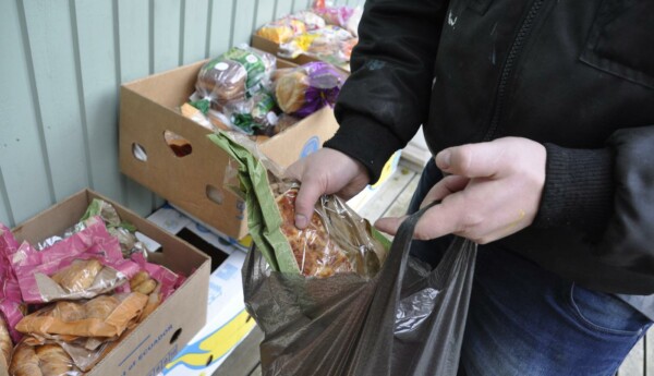 Händer som håller i en plastkasse med mat. Bakom syns kartonger fyllda med bröd