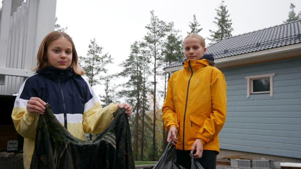 två flickor poserar med skräp de samlat: en mossig jacka och en stor sopsäck