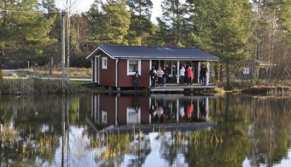 En liten stuga, med folk på terrassen, syns på andra sidan av en damm.