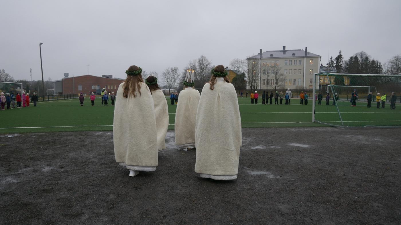 ett litet luciatåg bakifrån sett, går mot en sportplan i grått väder