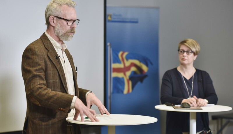 En man iklädd brun kostym står framför ett bord och talar. I bakgrunden syns en kvinna, samt en plansch med Ålands flagga.