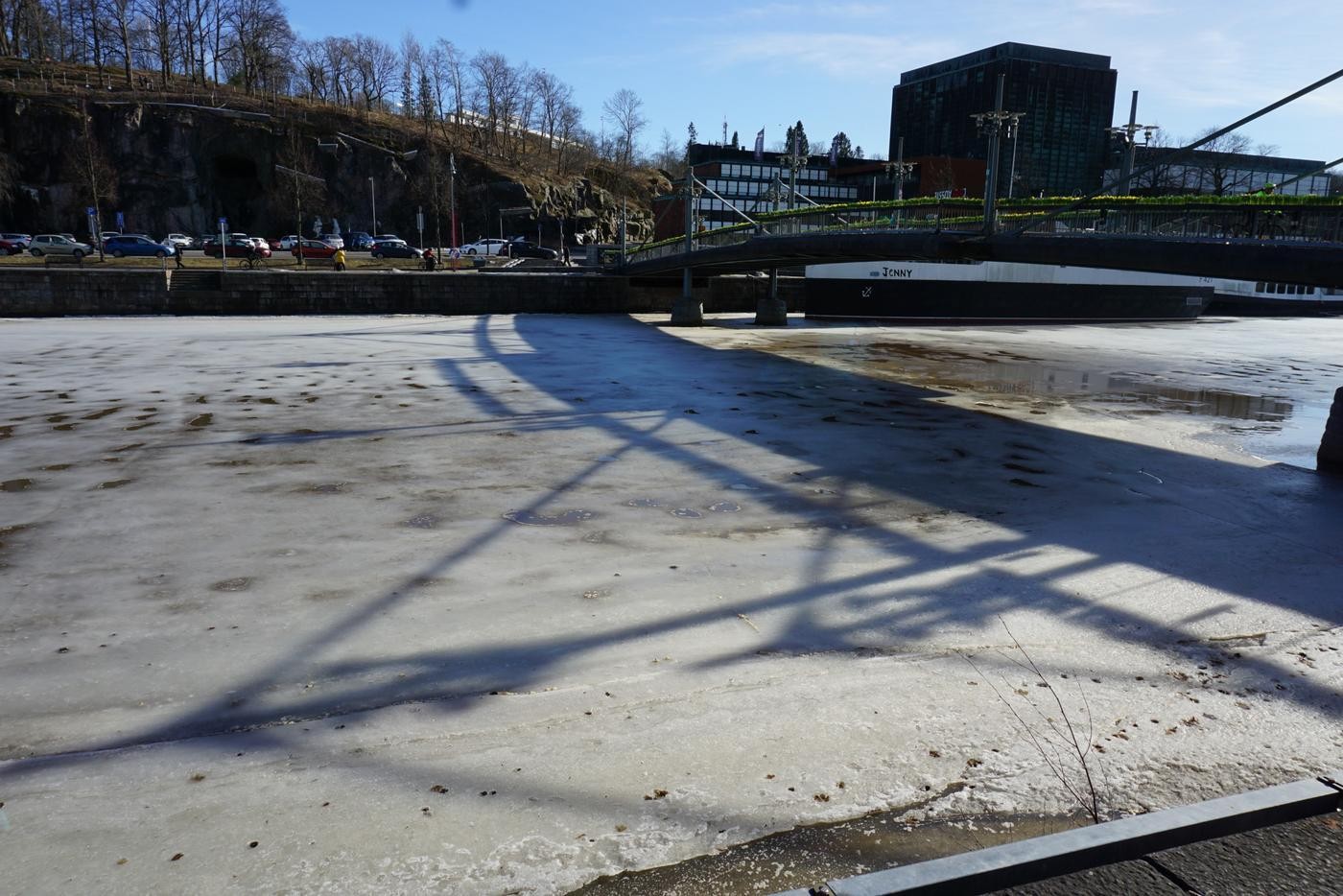 en gångbro kastar sin skugga över en delvis isbelagd å i tidig vårsol