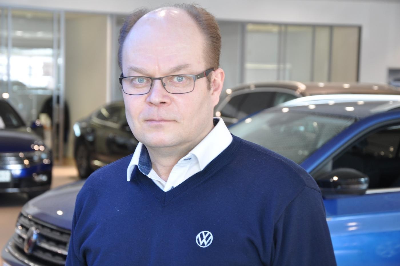 Bilförsäljare klädd i vit skjorta och blå tröja står framför bilar i en bilaffär