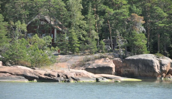 en sommarstuga skymtar bakom träden invid en strand med klippor