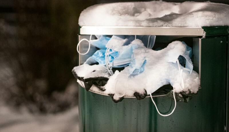 närbild: soptunna fylld med munskydd, täckt av snö.
