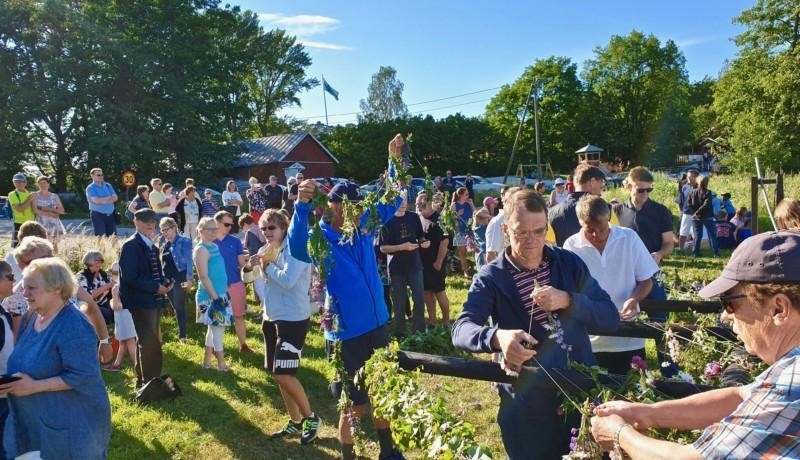 En grupp med människor varav en del är sysselsatt med att pryda midsommarstången som inte ännu är rest. Solig dag.