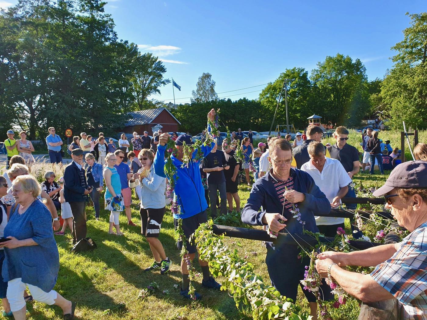 En grupp med människor varav en del är sysselsatt med att pryda midsommarstången som inte ännu är rest. Solig dag.