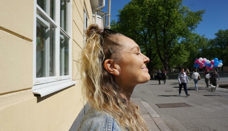 leende kvinna står vid en gul tegelvägg och blundar mot solen