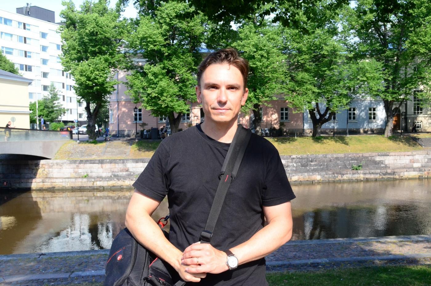 Man iklädd svart t-skjorta och blåa jeans står framför Aura å och tittar rakt in i kameran.