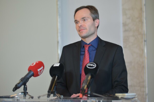 En man iklädd kostym står framför en rad mikrofoner och talar.