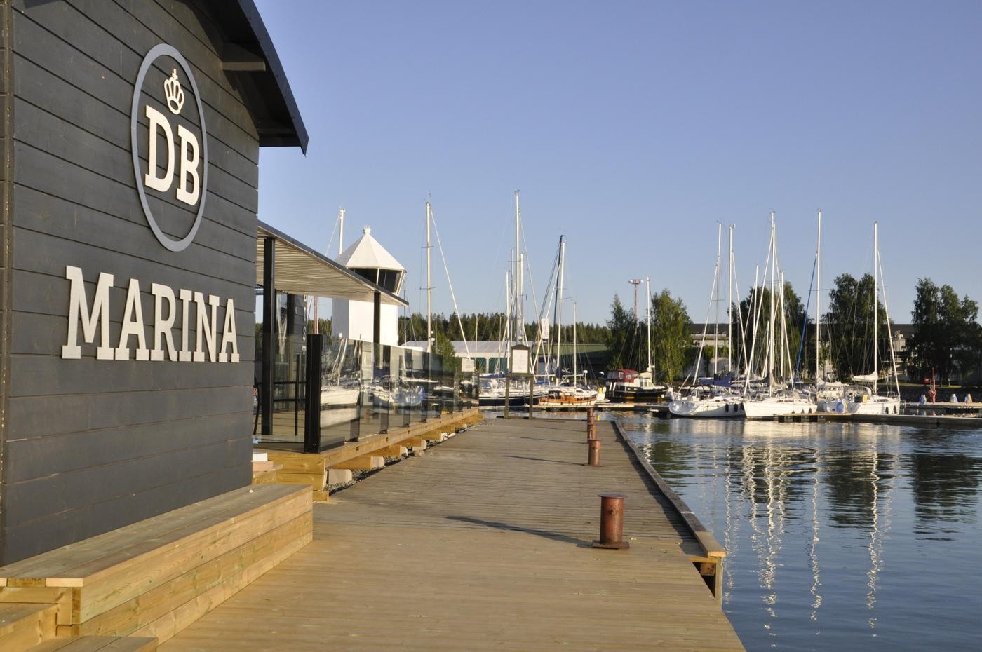 Lång brygga, vatten, segelbåtar i bakgrunden, svart byggnad till vänster med texten DB Marina på väggen.