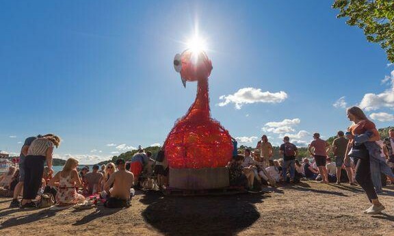 Ett stort konstverk i form av en flamingo står på en strand. Runt omkring sitter folk.