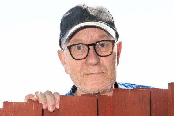 man med glasögon och keps tittar fram bakåt ett staket