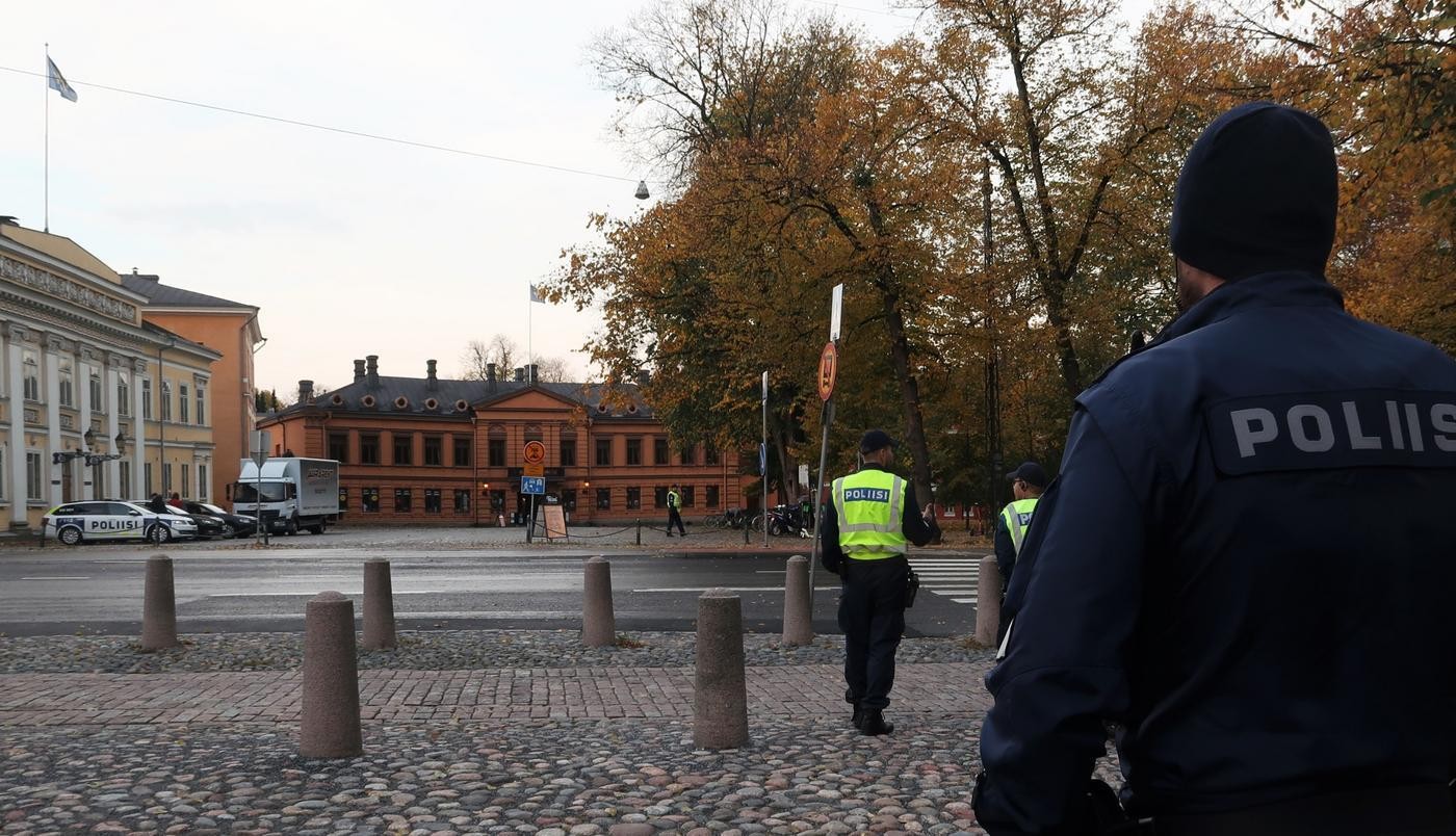 Bild av ryggen på en polis med blå polisjacka. Framför polisen syns ryggen av två poliser med neongul polisväst. På andra sidan vägen står en polisbilparkerad.