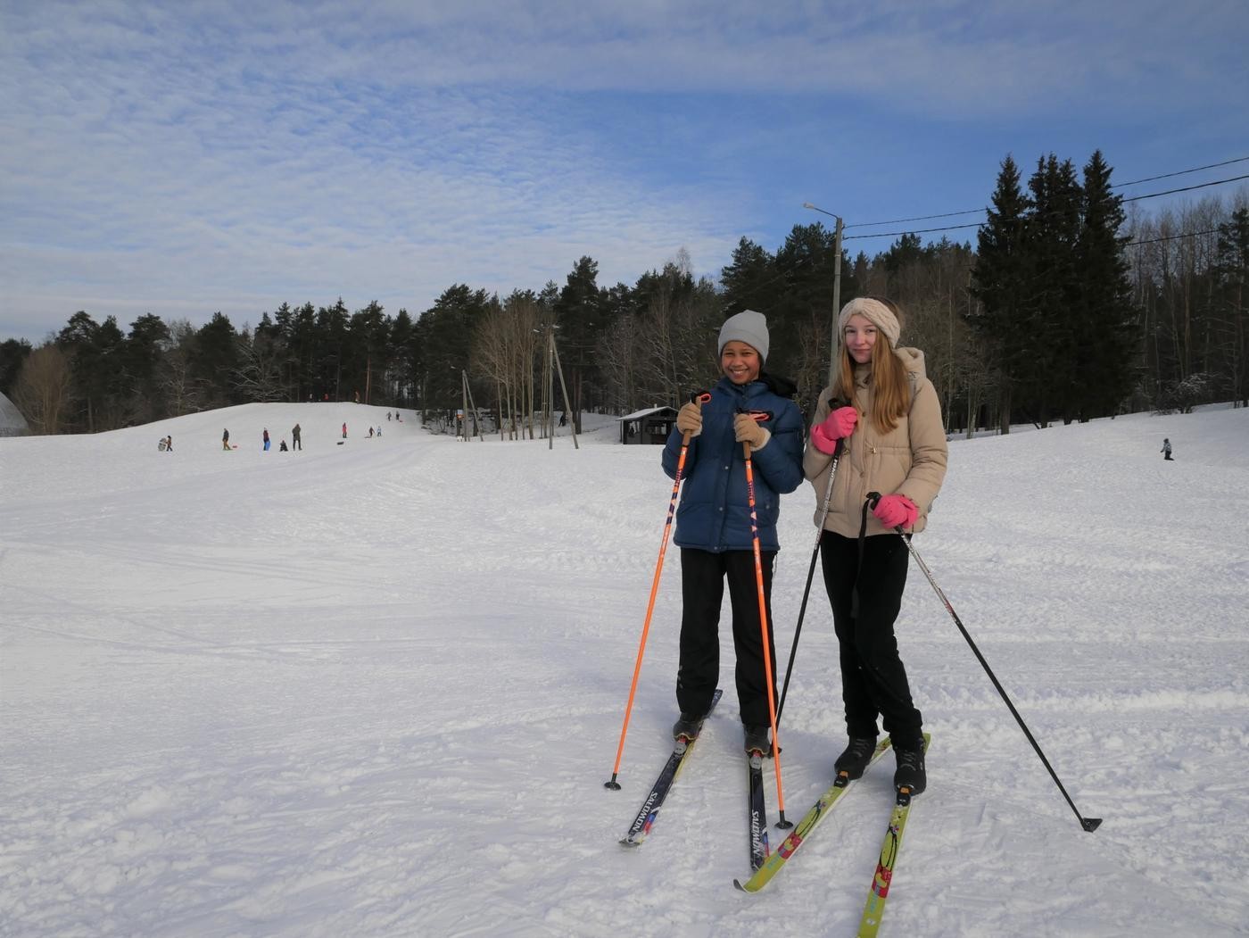 Två tjejer står på skidor och ler mot kameran. I bakgrunden skymtar skidspår och en pulkabacke.