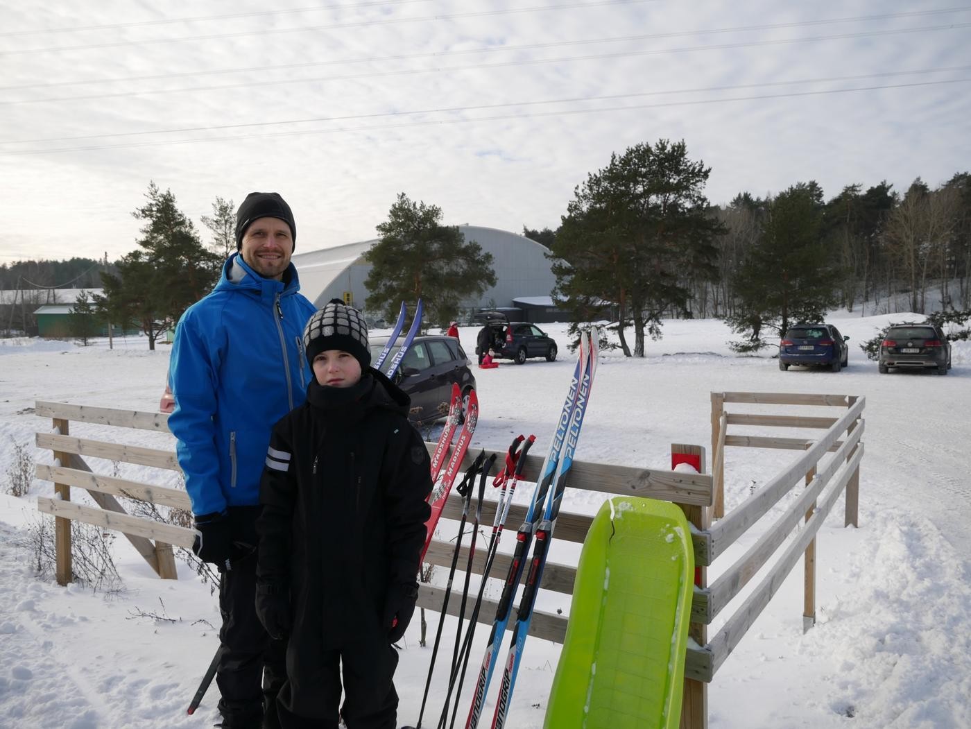 En man och en pojke ler mot kameran. Mot staketet bakom dem lutar skidor och en pulka.