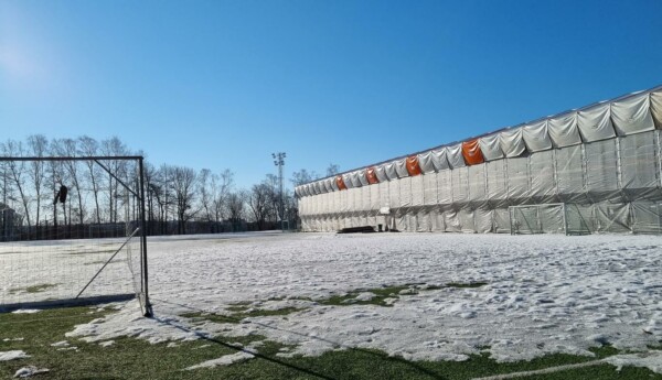 fotbollsplan med snö och inpaketerad läktare i bakgrunden, blå himmel