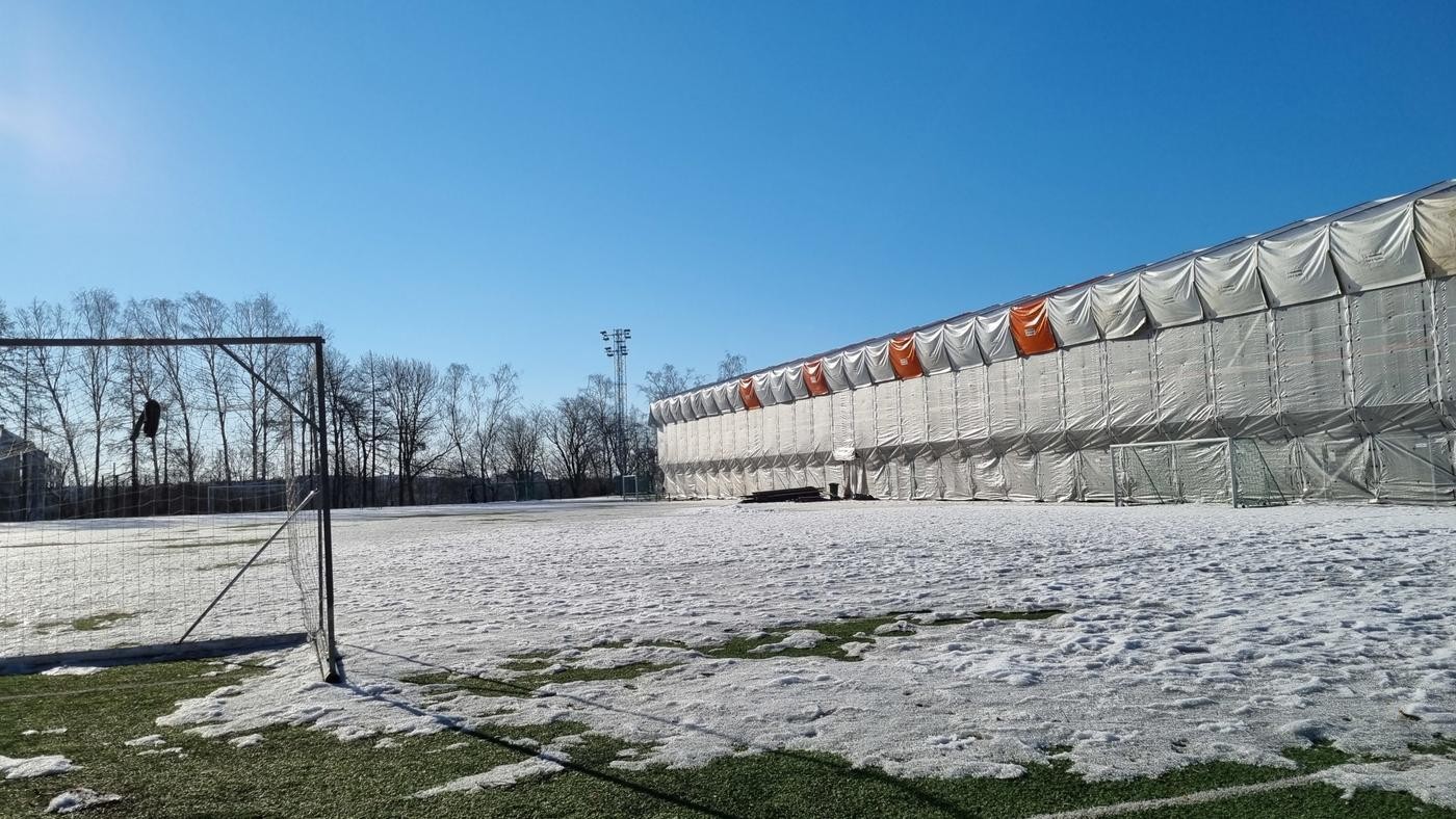 fotbollsplan med snö och inpaketerad läktare i bakgrunden, blå himmel