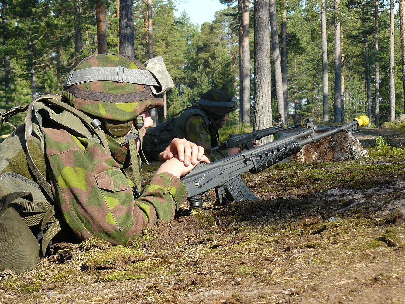 Två soldater med stormgevär ligger på marken i skogen